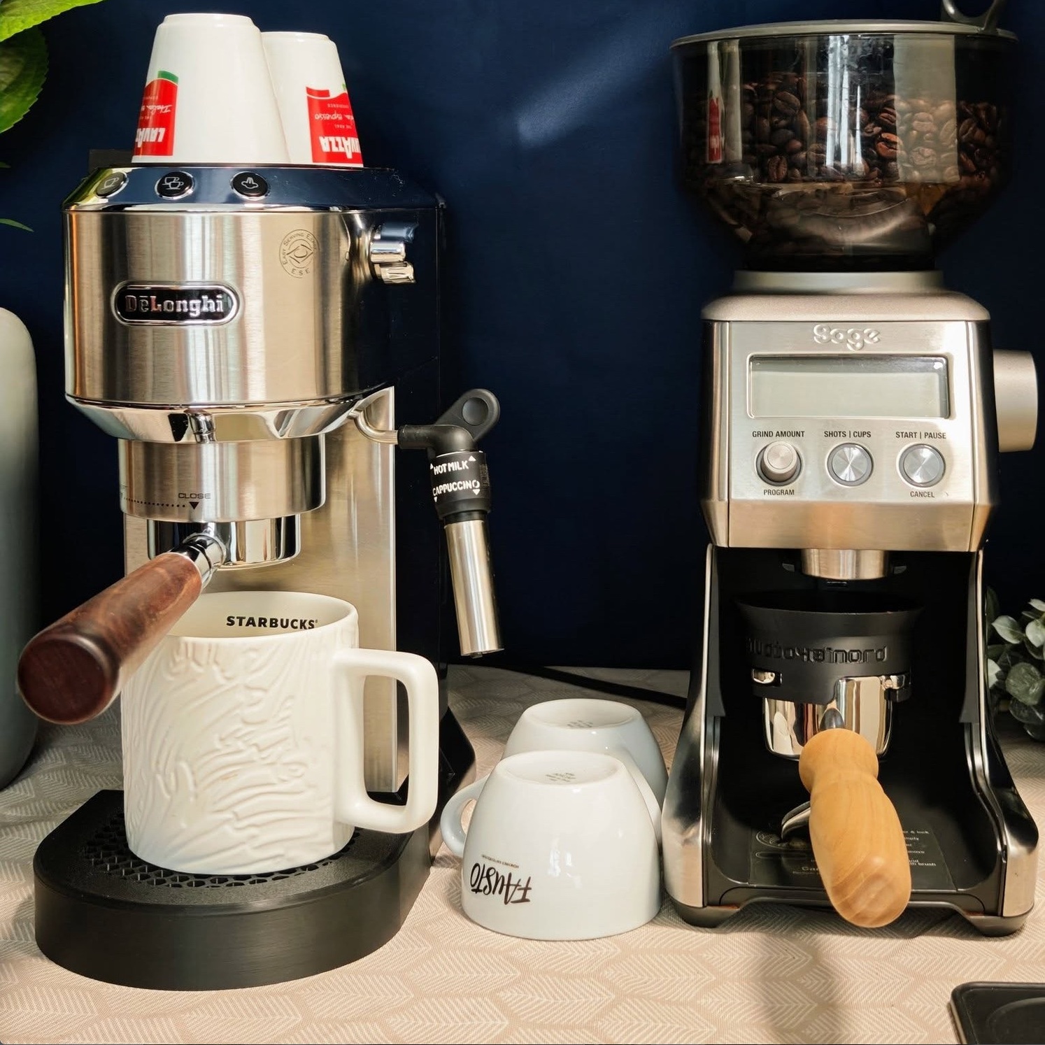 Silberne De'Longhi Dedica auf der Honeycomb-Abtropfschale, eine Tasse darunter, daneben eine Kaffeemühle mit eingesetztem Dosiertrichter, vor dunkelblauer Wand