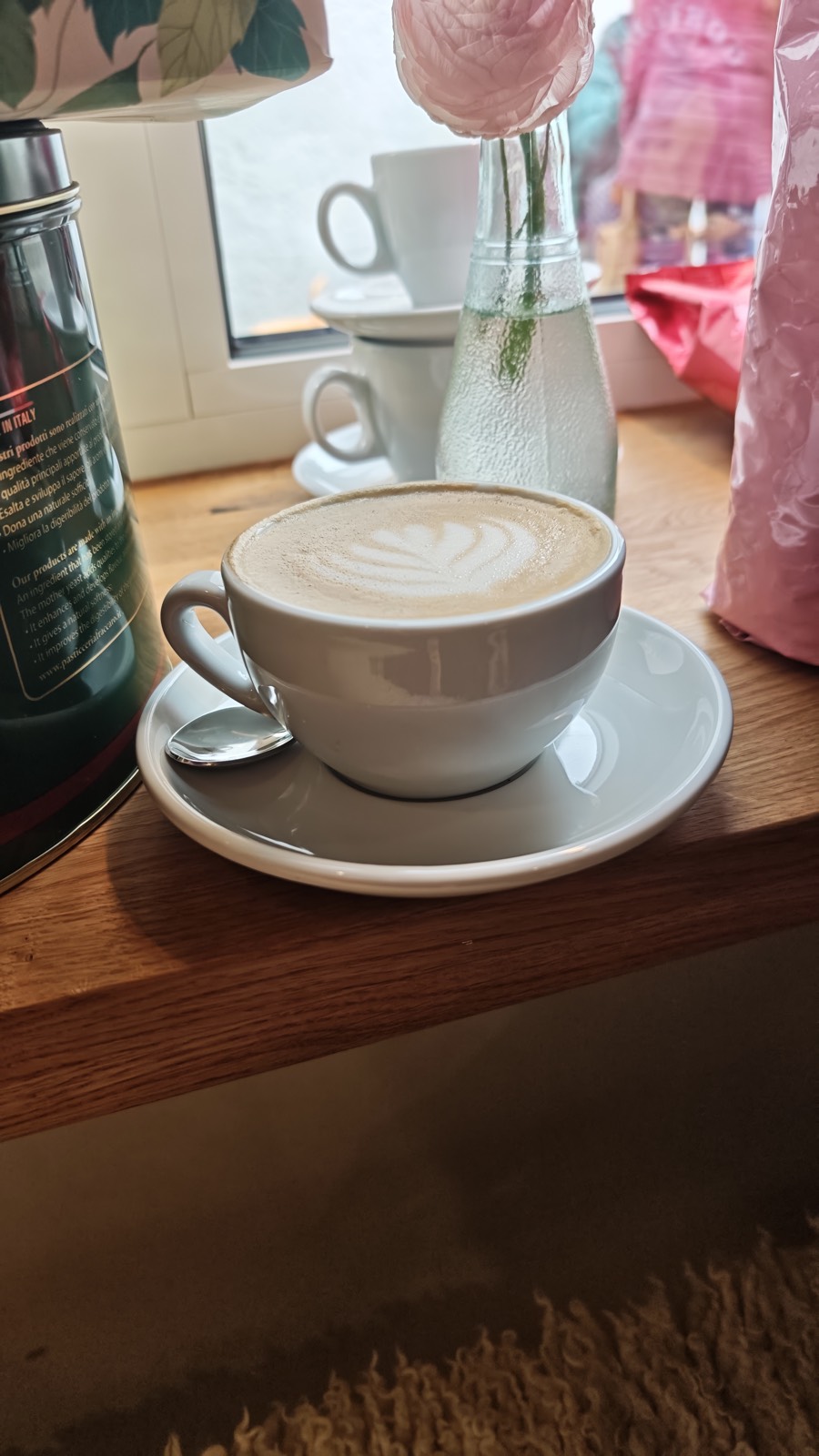 Cappuccino mit Latte-Art auf einer hölzernen Fensterablage, daneben eine Blume in einer Glasvase